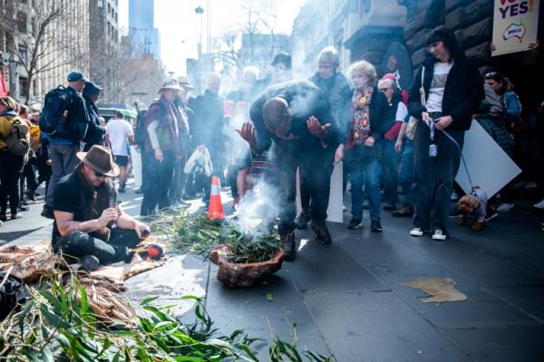 A man co<em></em>nducting a traditio<em></em>nal smoking ceremony on the road outside Melbourne Town Hall. He is leaning over a wooden bowl co<em></em>ntaining green leaves that are giving off smoke. A man to his side is speaking into a microphone. There are people watching the ceremony. 